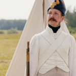 A historical reenactor in Napoleonic military uniform standing by a tent in open countryside.